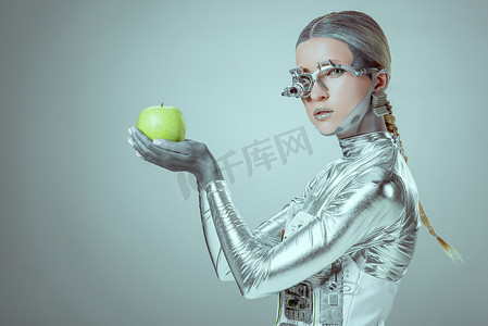 赛博格拿着绿色苹果的侧视图, 看着相机孤立的灰色, 未来的技术概念