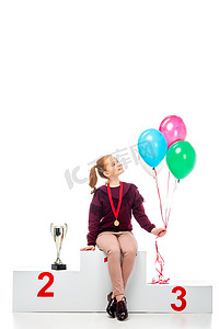 微笑的女学生坐在奖杯杯附近的冠军领奖台上, 拿着在白色孤立的五颜六色的气球