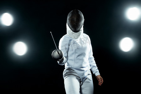 身穿击剑服装的年轻击剑运动员手持剑和面具。在黑色背景上与灯光隔离