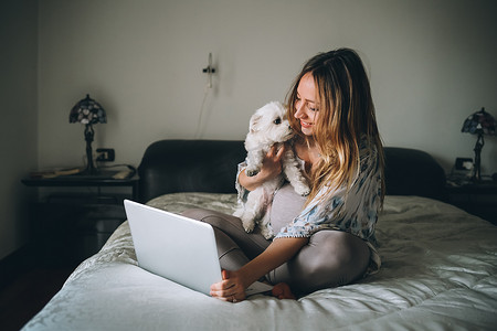 young woman indoors at home sitting bed using laptop computer holding dog - social network, wifi technology, connected online
