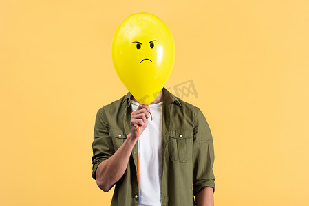 young man holding angry balloon in front of face, isolated on yellow