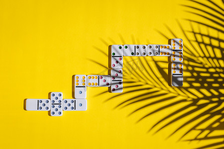 White dominoes with colorful dots lie like a snake on a yellow background with a shadow of a palm tree branch, a flat lay close-up.The concept of a summer board game.