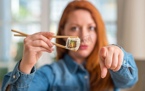 Redhead woman eating sushi using chopsticks pointing with finger to the camera and to you, hand sign, positive and confident gesture from the front