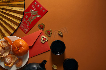 盘子上的橘子, 红茶, 带有象形文字的扇子, 金色的装饰和棕色背景上的红色信封的顶部视图
