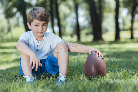 可爱的小男孩拿着橄榄球, 而坐在草地上, 看着相机