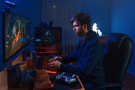 专注的职业游戏玩家游戏，参加在线竞赛，在家坐在电脑前。低光，霓虹灯色，侧视。专业的网络游戏玩家在电脑上进行在线游戏训练