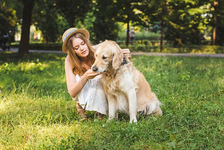 坐在草地摄影照片_白色连衣裙和草帽美丽的女孩在草地上坐在草地上看着狗的全长视图