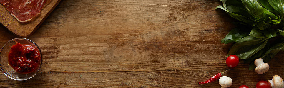 Top view of ingredients for pizza including tomato sauce, mushrooms, cherry tomatoes, chili peppers and prosciutto on wooden background, panoramic shot