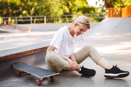 年轻的滑板女孩抱着她痛苦的腿与滑板附近的现代 skatepark