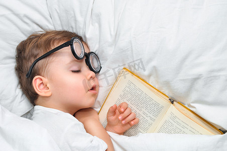 一个可爱的幼儿男孩正在戴着圆圆的眼镜睡在白色毯子下的旧书里.