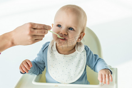 Mother with spoon feeding cute baby boy on feeding chair on white background