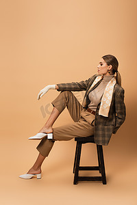 凳子上的摄影照片_穿着时髦西装和裤子的漂亮女人坐在米色背景的木制凳子上的侧视图