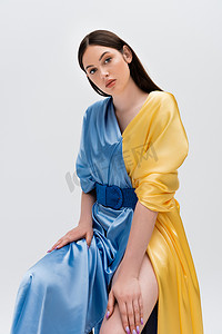 sensual ukrainian woman in blue and yellow dress posing while sitting isolated on grey