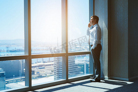 Mature and cnofident business executive looking looking out of large windows at a view of the city below, from the top floor of an office building, while talking on his mobile phone