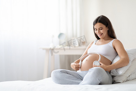 漂亮的孕妇穿着舒适的家居服，坐在床上，大肚子上拿着白色的无线耳机，笑着，复制空间。怀孕期间的音乐和婴儿发育概念