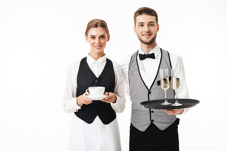年轻的微笑的侍者拿着杯子香槟, 而漂亮的女招待拿着杯咖啡在手愉快地看着在相机一起在白色背景