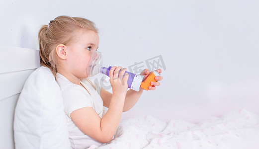 年轻病摄影照片_生病的小女孩躺在床上躺着吃哮喘药。有卧床吸入器治疗咳嗽的病童。流感季节。为年轻病人提供的卧室或医院房间。保健和药物治疗