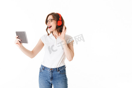 年轻快乐的女士在眼镜和耳机站立和愉快地看在白色背景的平板电脑的画像