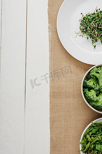 米黄色的盘子摄影照片_白色木制表面上的米黄色餐巾纸上的绿色蔬菜碗旁边盘子里新鲜的微绿色的顶部视图
