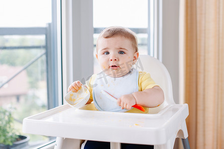 可爱可爱的白人孩子的肖像, 肮脏的凌乱的脸坐在高椅吃苹果泥与勺子。每天家庭的童年生活。婴儿尝试补充婴儿食品