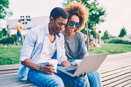 年轻积极的美国黑人夫妇身着时髦的衣服重新创造户外和观看有趣的视频网站上的笔记本电脑连接到4g。快乐的男人和女人在外面休息与小工具