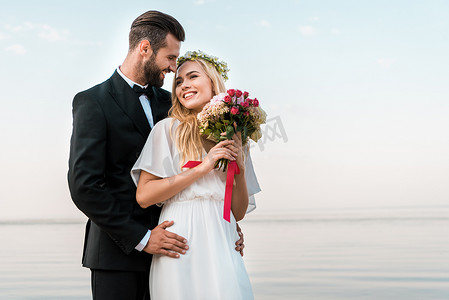 新郎拥抱微笑的新娘和她举行婚礼花束在海滩上