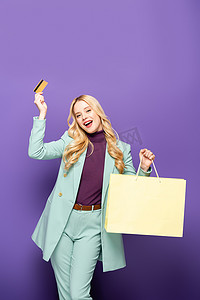 穿着时髦的蓝绿色西装、头戴信用卡、头戴紫色背景购物袋的快乐的金发女青年