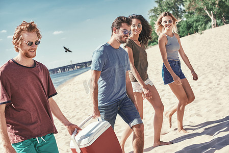 海滩上的朋友。后视图的欢快的年轻人走在海边的海滩, 而两名男子携带塑料冷却器
