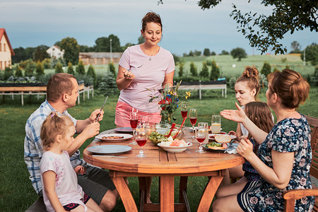 外出野餐摄影照片_夏天外出野餐时,一家人在自家花园的烤架边吃饭.围坐在餐桌旁，端着食物和碗碟的人
