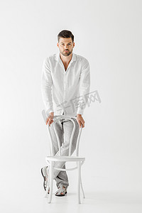 年轻男子穿着亚麻衣服站在附近的椅子上孤立的灰色背景 