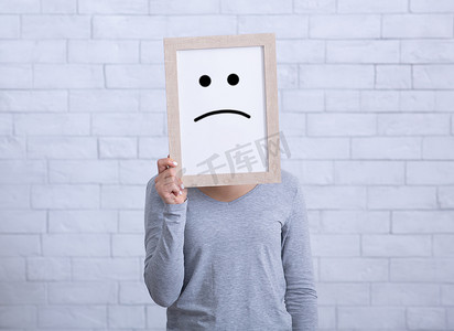 lady摄影照片_Millennial lady holding picture frame with unhappy emoticon, covering her face against white wall