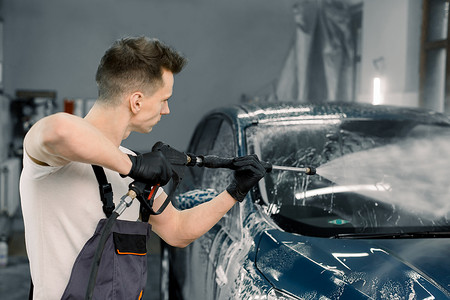 新店开业洗车摄影照片_一个工人在洗车时用高压水清洗现代蓝色汽车的侧角视图。洗车服务及详细概念