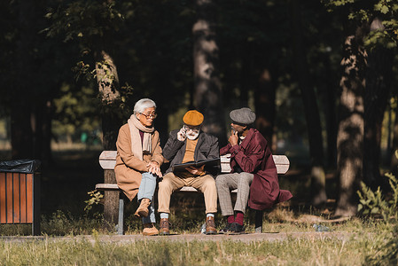 资深跨种族人士坐在好友身边，带着相册，在公园里用智能手机交谈 