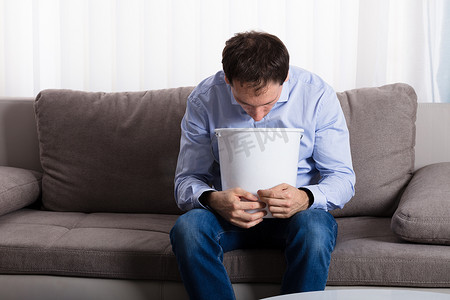一个男人坐在沙发上呕吐在家里的白色塑料桶