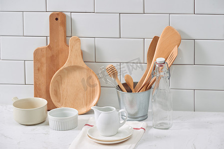 白木墙简单质朴厨具 ︰ 粗陶瓷壶与木制烹饪器具套、 成堆的陶瓷碗、 壶、 木托盘.