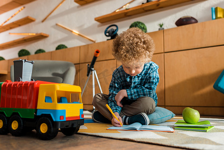 聪明的孩子坐在靠近苹果、玩具车和书本的地板上，一边在笔记本上写字 