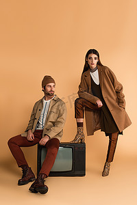秋装摄影照片_穿着时髦秋装的女人坐在米色背景的老式电视机前，看着镜头旁边的时髦男人