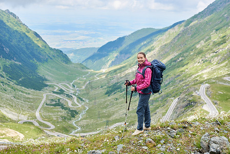 微笑的女性游客站在与步行坚持到美丽的景色华丽缠绕 Transfagarashan 路在罗马尼亚。女子登山者青山山快乐岩石旅游路线路径