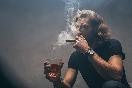 fashionable businessman in black outfit smoking cigar and drinking whiskey near grey wall