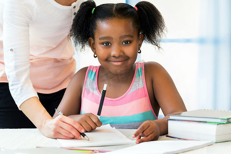 书桌纸摄影照片_关闭可爱的小非洲学生的肖像做家庭作业与白人教师。Ponytailed 的女孩坐在书桌与钢笔, 纸和书.