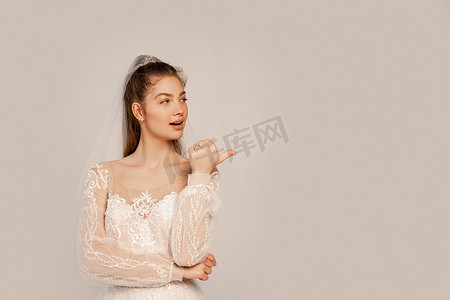 young bride looking away and pointing with thumb isolated on grey 