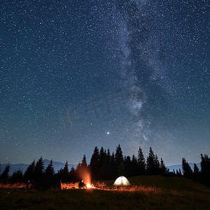 在美丽的夜空下，男性旅行者坐在帐篷边，望着篝火，点缀着星空和银河。旅行、远足和夜间露营的概念.