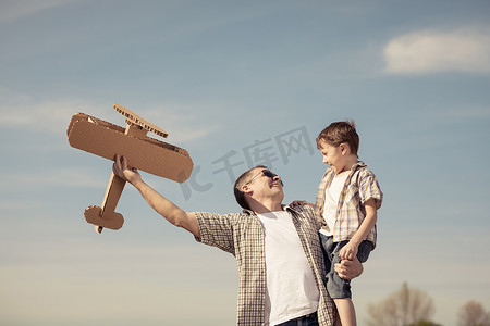 飞机里摄影照片_爸爸和儿子白天在公园里玩纸板玩具飞机。友好家庭的概念。在蓝天的背景上拍摄的图片.