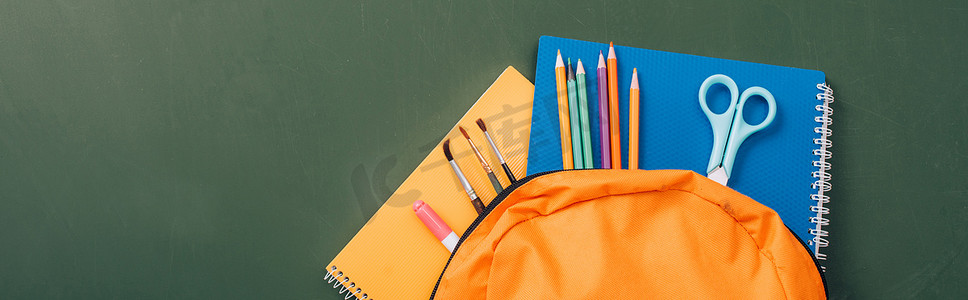 绿色黑板上黄色背包中笔记本、彩色铅笔、剪刀和画笔的水平图像