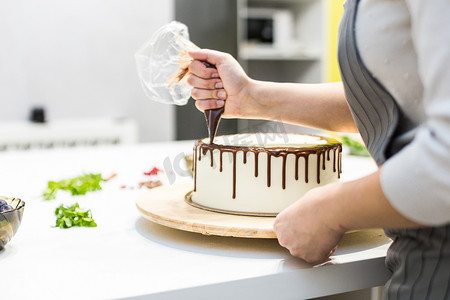 液体巧克力摄影照片_糖果师将水果袋中的液体巧克力挤压到木制支架上的白色奶油饼干蛋糕上。自制糕点、烹饪蛋糕的概念.