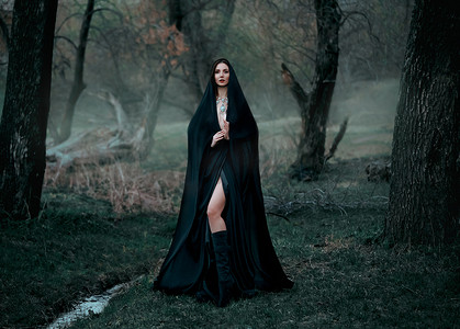 神秘的幻想哥特式女人被邪恶缠住了。穿着黑衣斗篷的女恶魔吸血鬼。走在密密麻麻的森林深处，树木丛生。中古女王穿著丝绸斗篷，围巾摆姿势