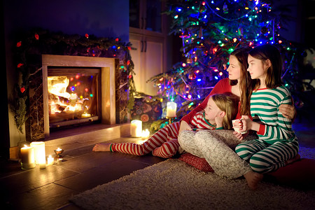 平安夜，快乐的年轻妈妈和她的女儿们一起坐在一个温暖的黑暗客厅的壁炉边，度过了一个愉快的夜晚。 在家里庆祝圣诞节.