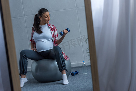 镜子与反射的非洲裔美国人怀孕的妇女坐在适合球和训练与哑铃