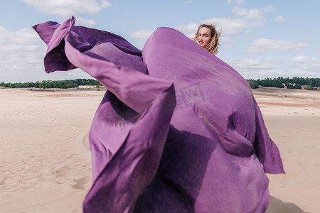 紫色衣服的女孩摄影照片_一个穿着米色衣服，手里拿着紫色布的年轻纤细姑娘，在沙漠中迎风而坐