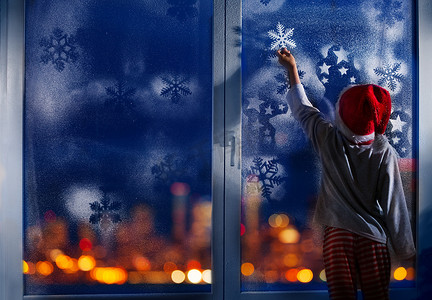 圣诞老人帽子摄影照片_圣诞前夜, 戴着圣诞老人帽子的小男孩在冰冷的窗户上触摸雪花 